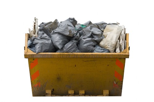 A large yellow industrial skip filled with various rubbish bags and waste materials, positioned on a paved surface outdoors. The waste bags are predominantly grey, made of plastic, and appear to be tightly packed and stacked within the skip. There is a single white bag among the grey ones, creating contrast. The skip’s exterior shows signs of dirt and mild wear, with red and orange reflective stripes on each side providing visibility. The scene is set in a utilitarian environment, typical of waste removal sites, possibly along a driveway or a service area in Alperton. The image emphasizes the collection or disposal of commercial waste, aligning with the services offered by Commercial Waste Alperton for rubbish removal.