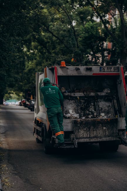 A worker wearing a green uniform and safety helmet is positioned beside a small commercial waste collection vehicle on a quiet urban street lined with trees. The vehicle's rear hatch is open, revealin